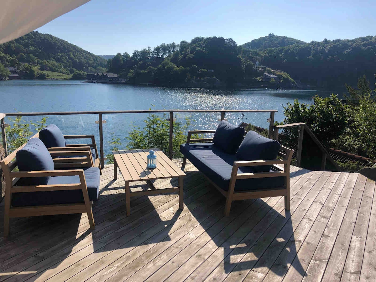 A wooden deck overlooks a serene body of water, where gentle ripples reflect the surrounding green hills. Comfortable seating in navy blue cushions is arranged around a small table, inviting relaxation while enjoying the tranquil views.