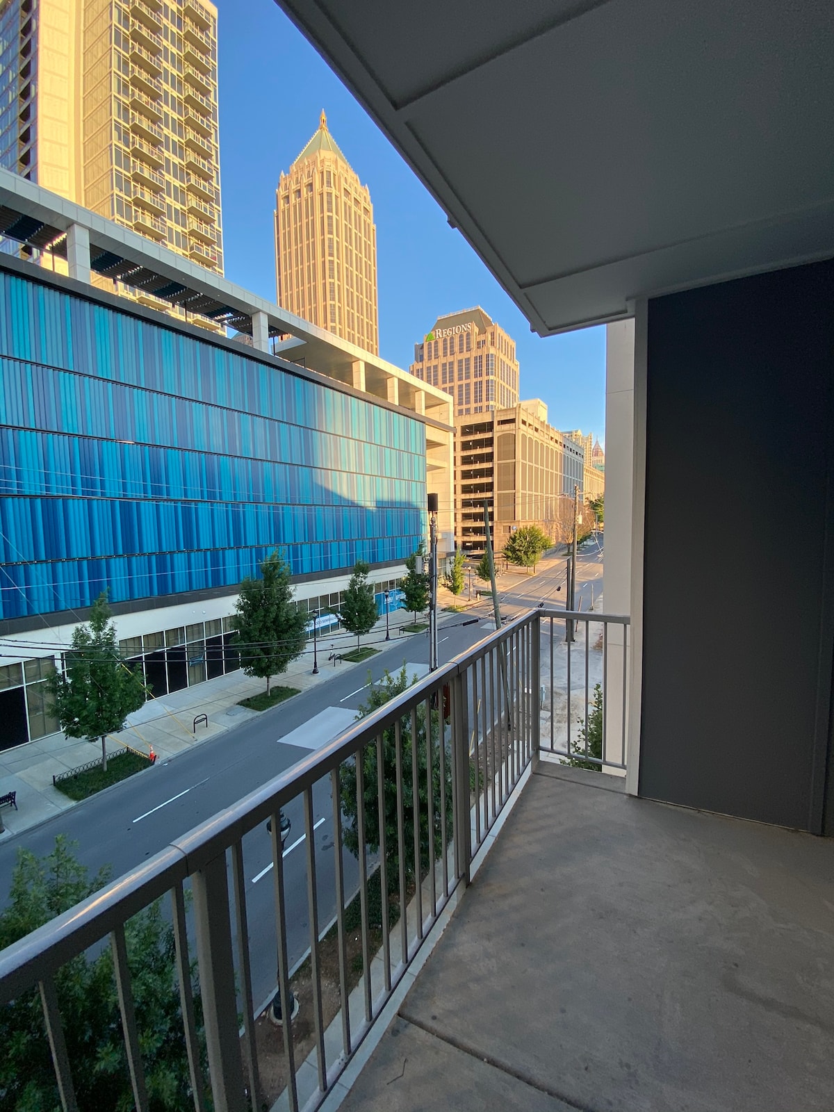 A balcony offers a view of the city skyline, showcasing modern high-rise buildings and a clear blue sky. Sidewalks lined with trees are visible below, contributing to the vibrant urban atmosphere.