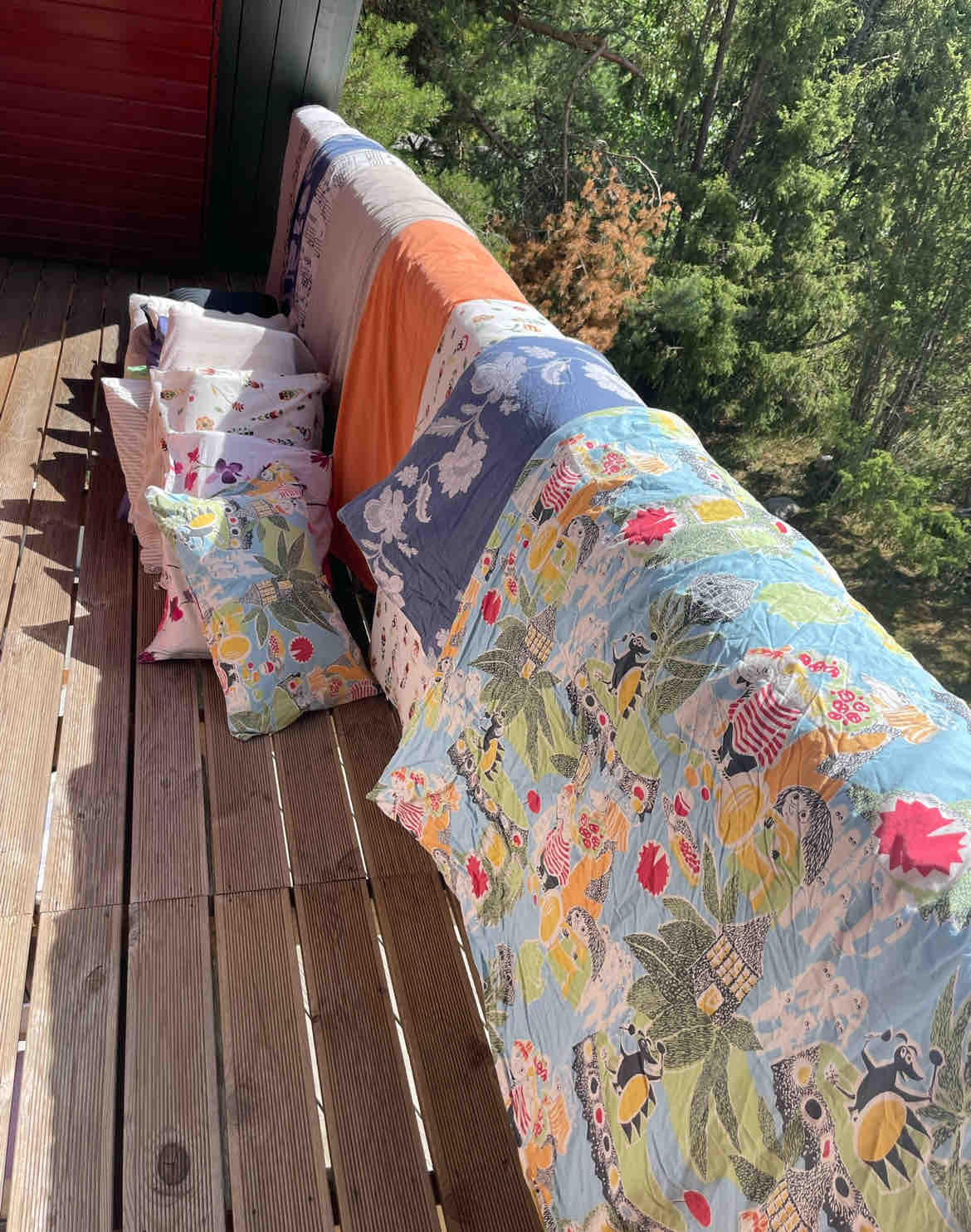 A collection of decorative pillows and quilts adorns the wooden outdoor seating area. Bright floral patterns and a variety of colors are displayed, complementing the natural surroundings. Sunlight filters through the trees, creating a warm ambiance on the deck.