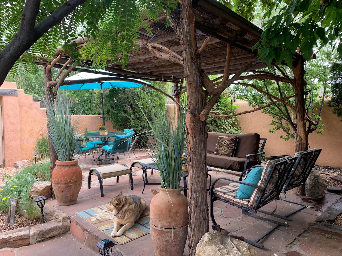 A shaded outdoor seating area is highlighted by a wooden pergola, offering a comfortable sofa and several chairs arranged around a stone patio. Potted plants and decorative stones enhance the peaceful garden setting, with an umbrella providing additional shade for a nearby dining table.