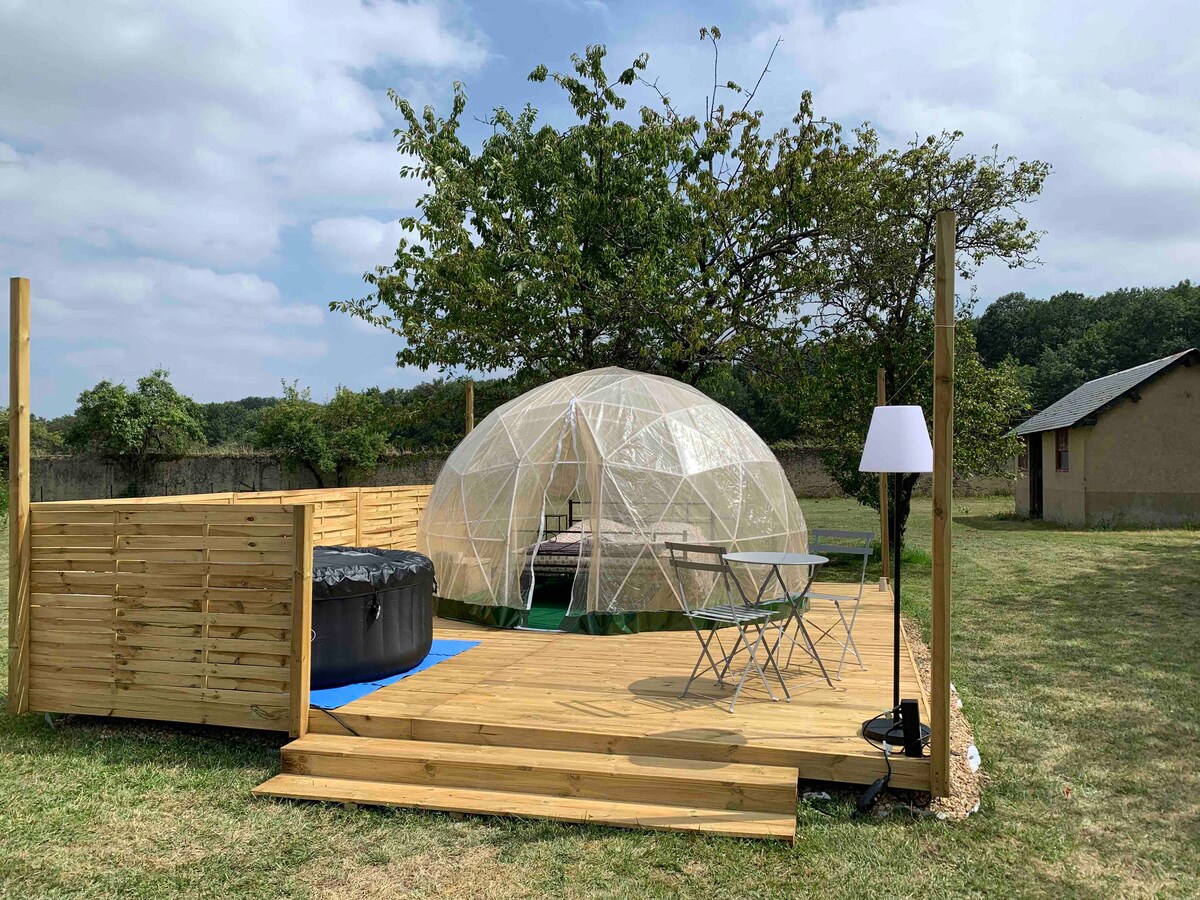 A dome structure surrounded by a spacious wooden deck is set in a grassy area. In front of the dome, a circular hot tub is placed. A small dining table with two chairs is positioned nearby, accompanied by a modern floor lamp. A tree provides partial shade.