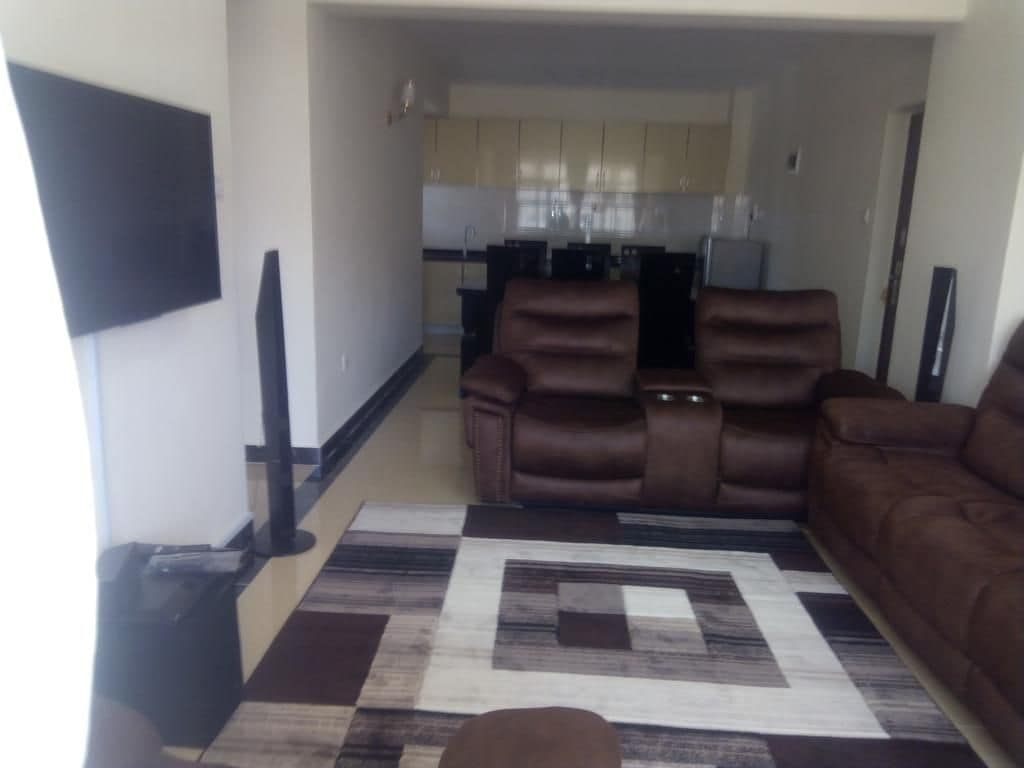 A living area showcases a large, plush brown sofa set arranged around a patterned area rug. A mounted flat-screen television is visible on one wall, with a glimpse of the open kitchen area containing modern cabinetry and appliances in the background.