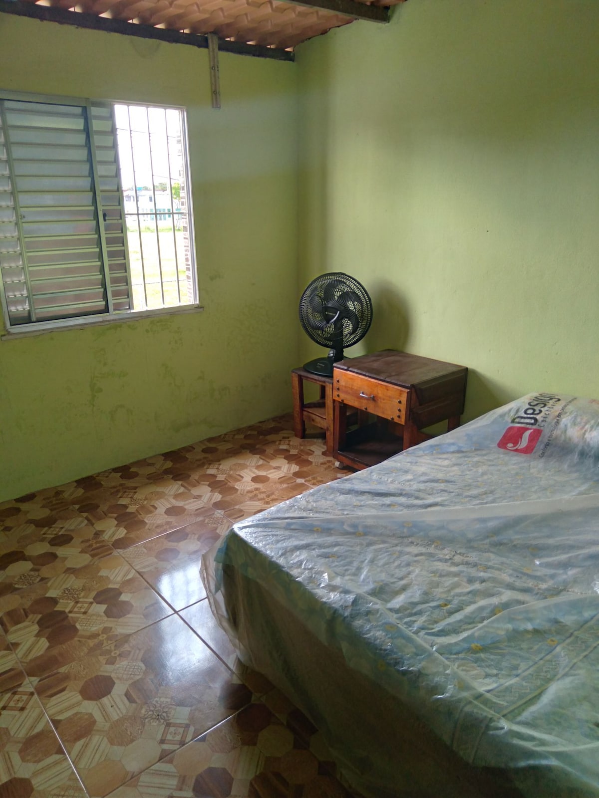 A room features a spacious bed covered with a protective plastic sheet. A wooden nightstand sits beside the bed, and a fan is placed nearby. Natural light enters through a window with metal grilles, illuminating the patterned tiled floor.