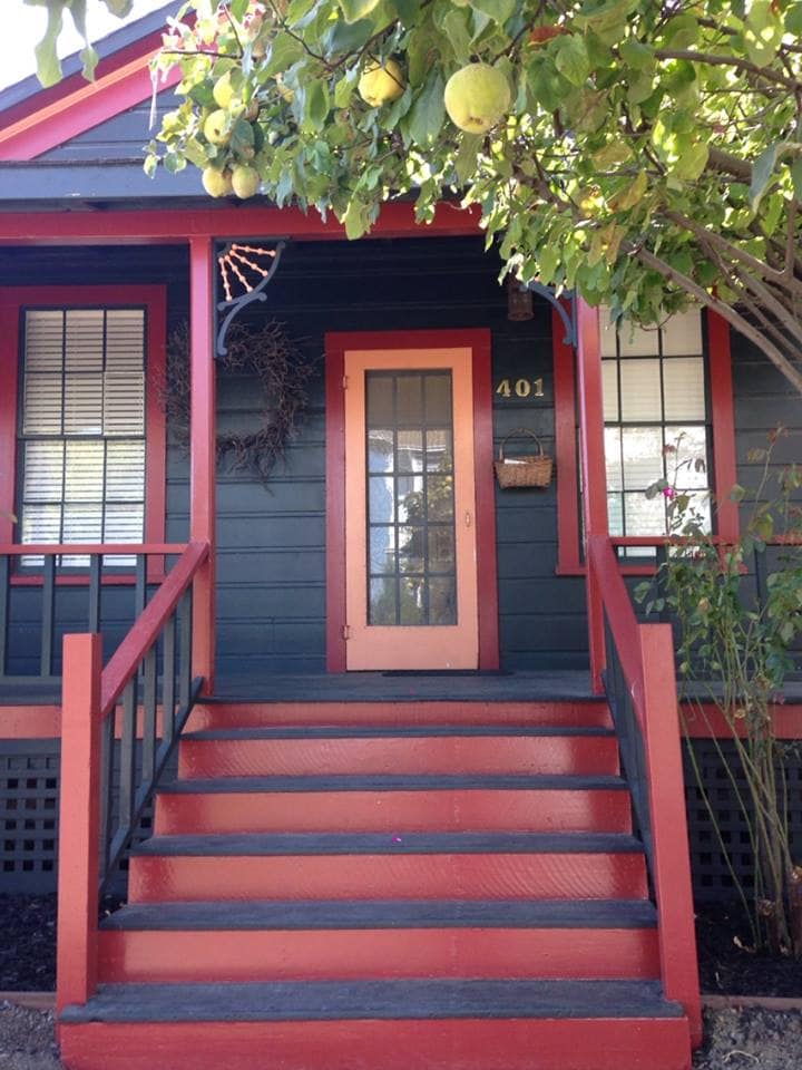 The entrance features a vibrant red staircase leading to a door with glass panels and decorative frame. The exterior is painted in dark hues, accented by a small window beside the door. A tree with yellow fruit is seen above, adding a natural touch.