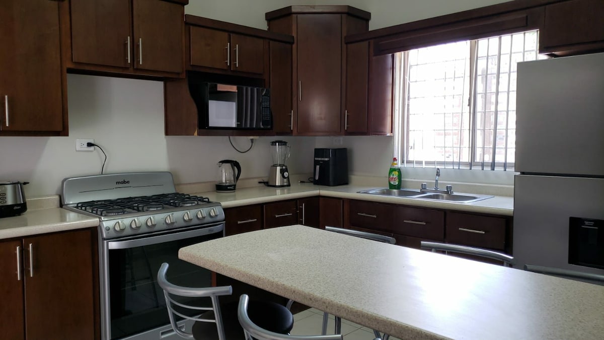A modern kitchen features dark wood cabinetry and stainless-steel appliances. A spacious island provides seating, and a large window allows natural light to brighten the room. Essential appliances are visible, including a gas stove, microwave, and refrigerator.