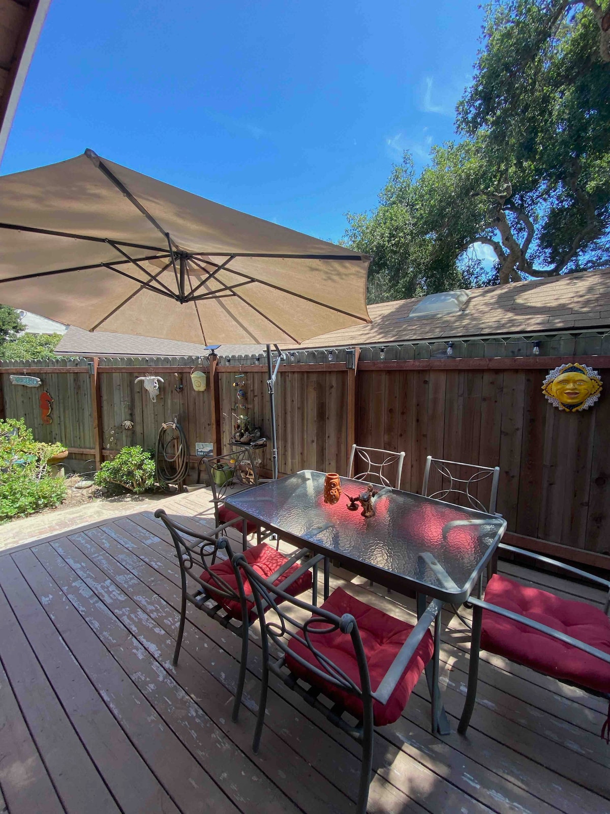 An outdoor dining area is presented on a wooden deck, furnished with a glass-top table surrounded by four metal chairs with red cushions. A large umbrella provides shade overhead, and greenery surrounds the space, enhancing the private and tranquil setting.