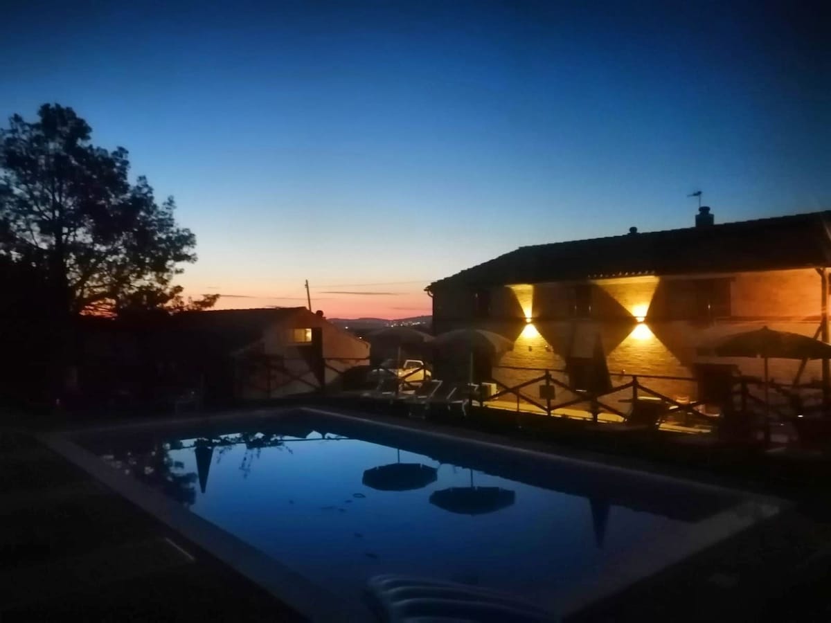 The scene captures a tranquil outdoor pool area illuminated by soft lighting. A two-story building is visible, featuring warm lighting that enhances the evening atmosphere. The sky transitions from deep blue to orange, indicating sunset, while silhouettes of trees frame the pool.