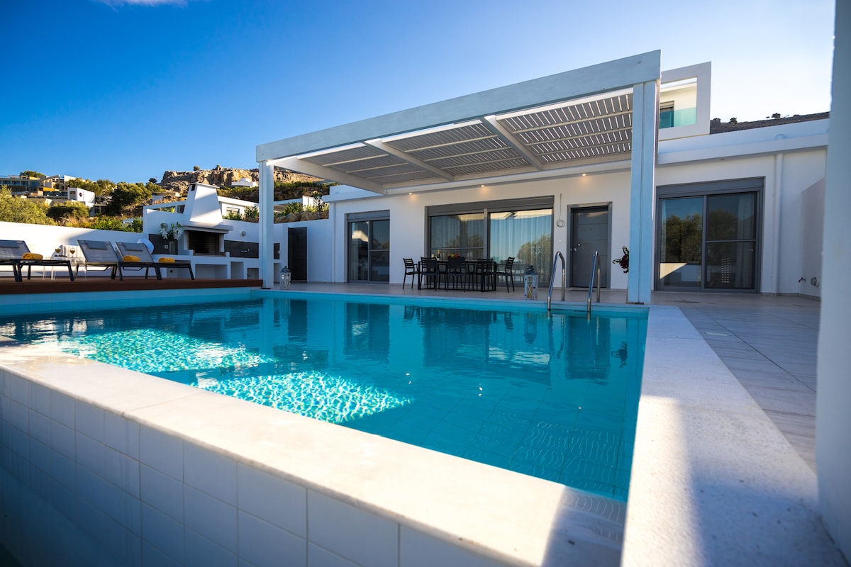 A modern villa is depicted with an infinity pool reflecting the clear blue sky. The outdoor dining area is visible under a shaded pergola, with several lounge chairs positioned nearby. Large glass doors lead into the villa, enhancing the open layout.