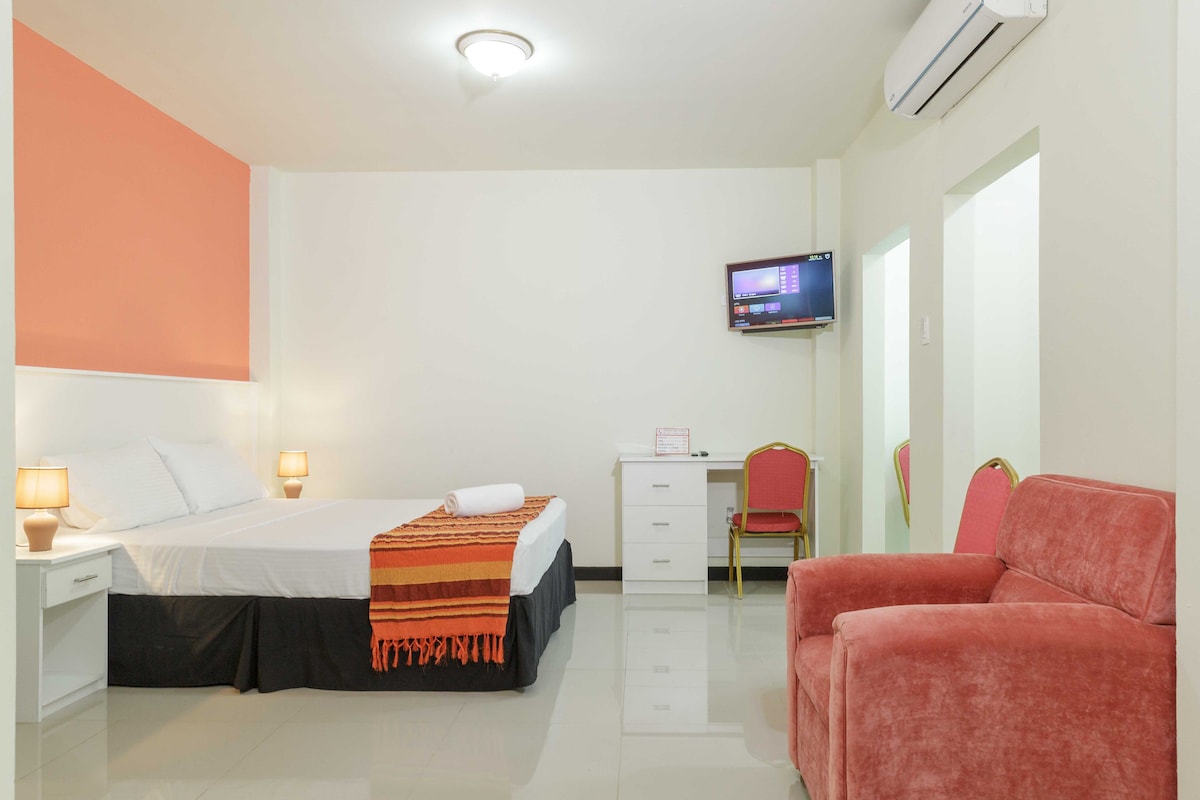 A comfortable room features a spacious bed with a colorful throw blanket. Two bedside lamps provide soft lighting. A small TV is mounted on the wall, and a dresser is positioned beside a pair of vibrant chairs. The polished floor reflects the bright walls.