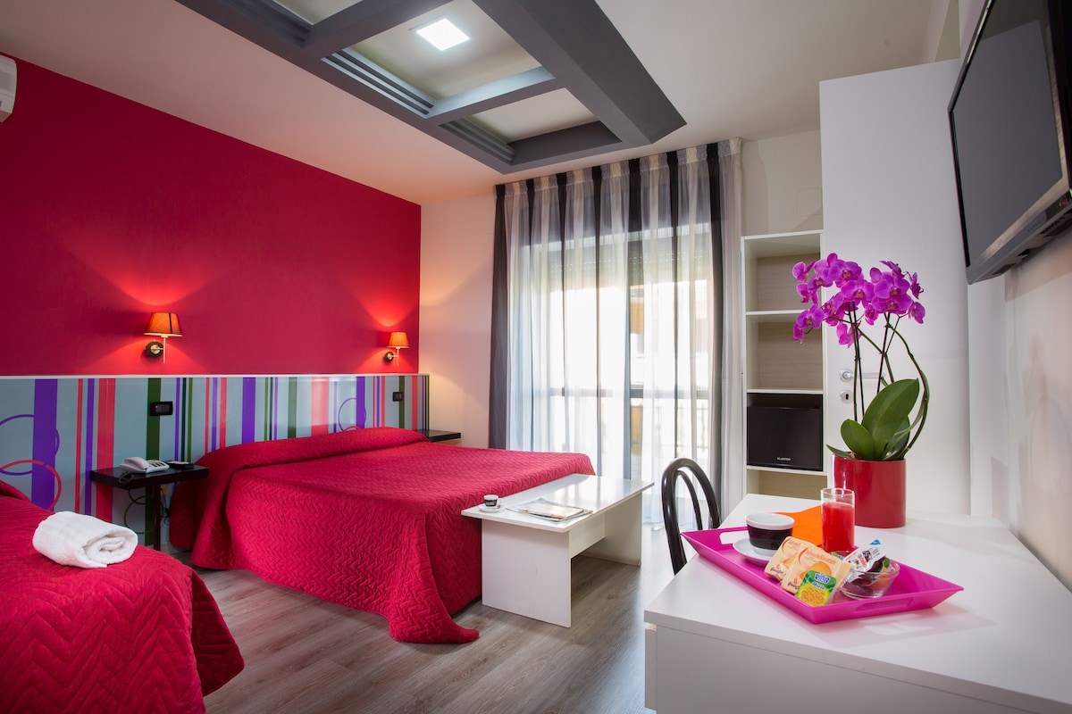 A well-appointed guest room features a vibrant red color scheme with striped accents. Two beds are adorned with matching red bedspreads. A small table holds refreshments, and a stylish orchid adds a touch of greenery. Natural light filters through sheer curtains, enhancing the inviting ambiance.