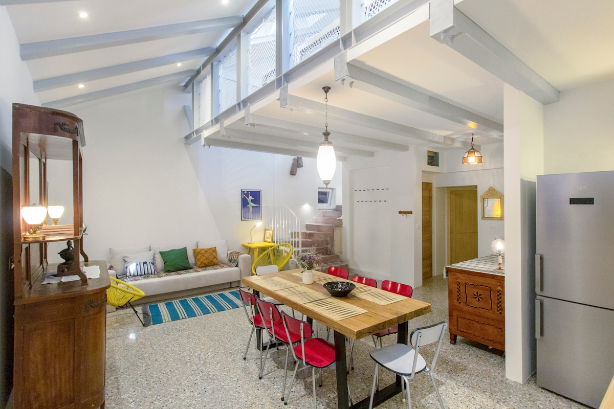 An open-plan living area features high ceilings and wooden beams, creating a bright and airy feel. A dining table made of wood is surrounded by colorful chairs. A comfortable sofa is positioned nearby, with decorative cushions adding a touch of color. A modern kitchen is visible in the background.