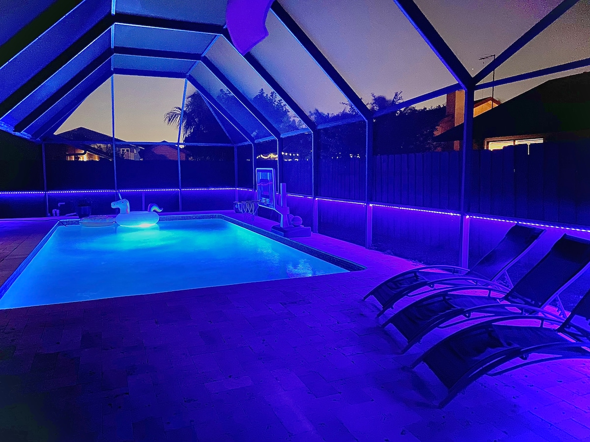 A heated pool area is enclosed by a screen, featuring ambient lighting that casts a soft blue glow. Lounge chairs are positioned beside the pool, inviting relaxation. The surrounding area includes a well-maintained patio and glimpses of the evening sky through the screened enclosure.