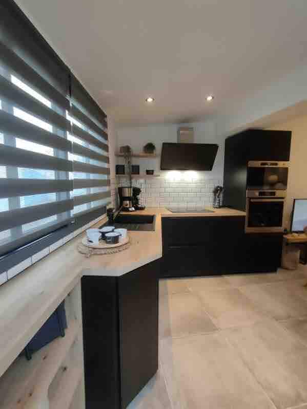 The kitchen features modern black cabinetry, a built-in oven, and a sleek cooktop. A coffee maker and other appliances are neatly arranged. A window with blackout shades allows natural light to illuminate the space, enhancing the clean, functional design.