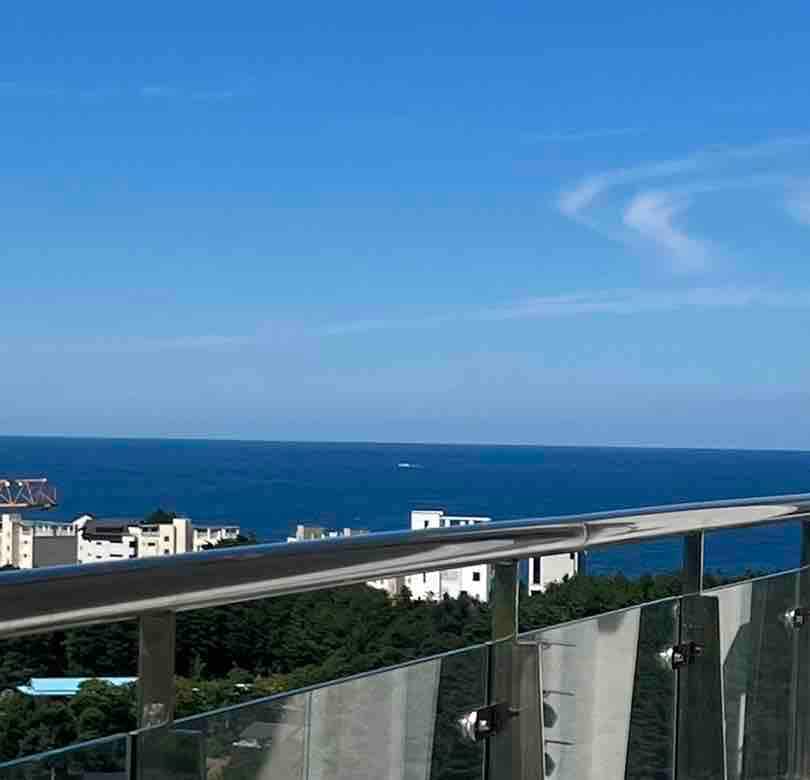 A spacious balcony overlooks the shimmering sea, framed by a glass railing that enhances the view. The clear sky extends into the horizon, with buildings nestled along the coastline creating a serene atmosphere.