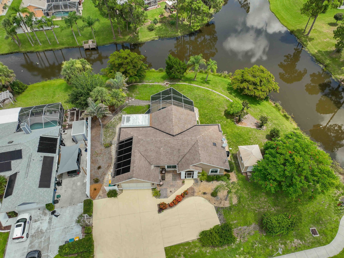 An aerial view captures the spacious home surrounded by lush greenery, with the canal visible at the rear. The large driveway leads to a well-maintained yard, highlighting the proximity to water and the beautifully landscaped surroundings.