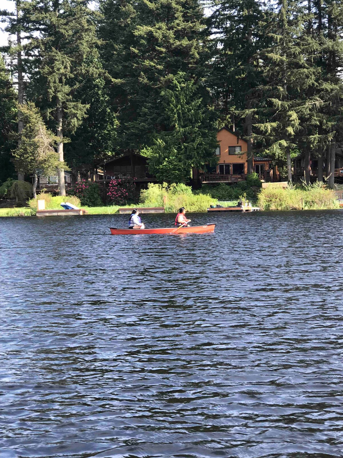 Two individuals are seated in a red canoe, gently paddling on a glistening lake. Lush green trees line the shore, contrasted by the wooden cabin in the background, which displays a warm color palette. The tranquil scene invites outdoor recreation amid a natural setting.