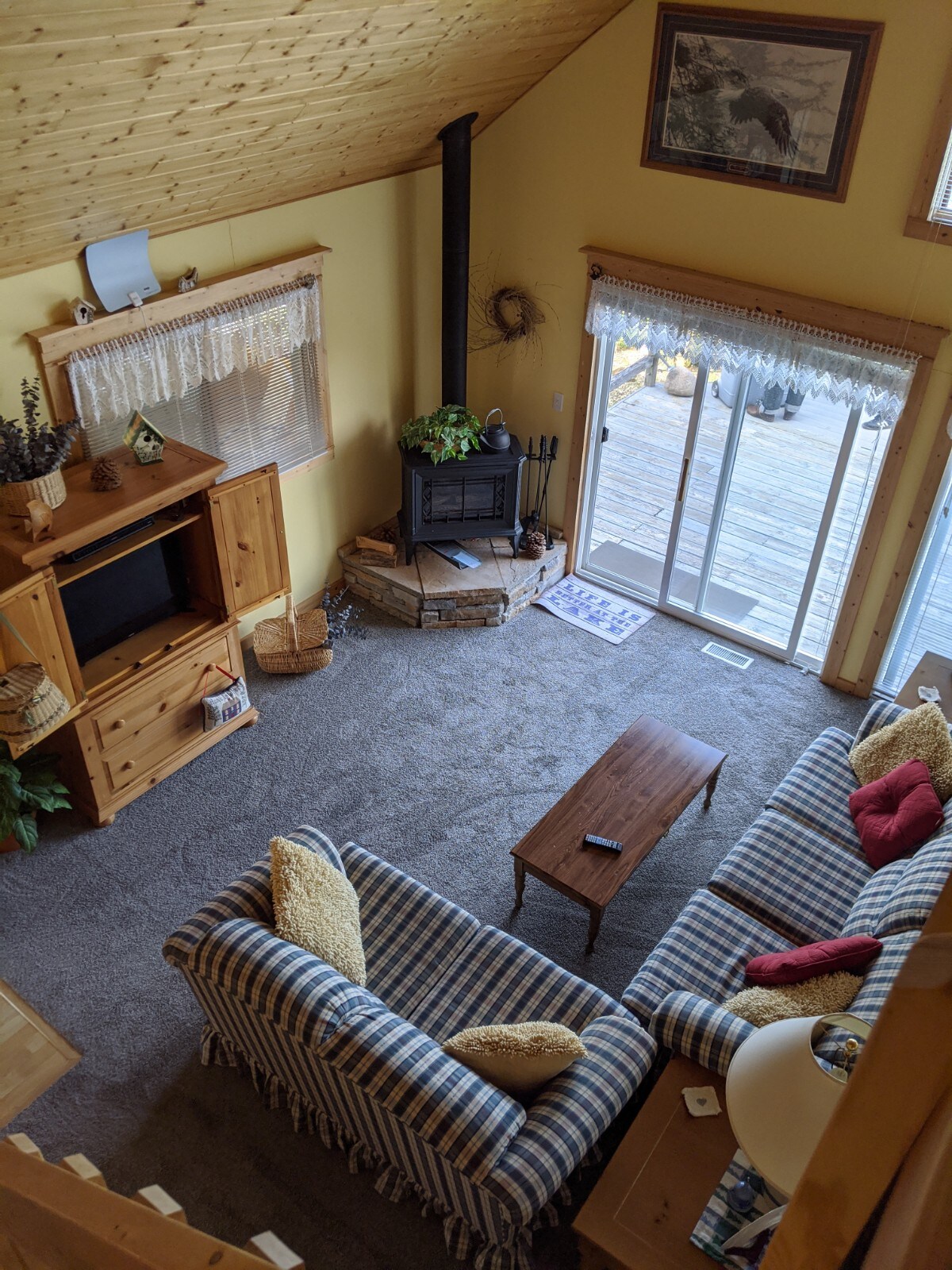 The inviting living area features a striped sofa and a cozy coffee table, with warm yellow walls and a wooden entertainment unit. A wood-burning stove provides warmth, while sliding glass doors lead to a deck, allowing natural light to fill the space.