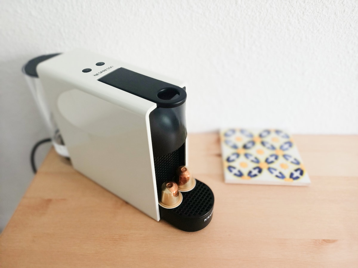 A sleek Nespresso coffee machine is positioned on a light wooden countertop, featuring two copper coffee capsules resting in the holder. A decorative coaster with colorful patterns is placed nearby, adding a touch of charm to the workspace.