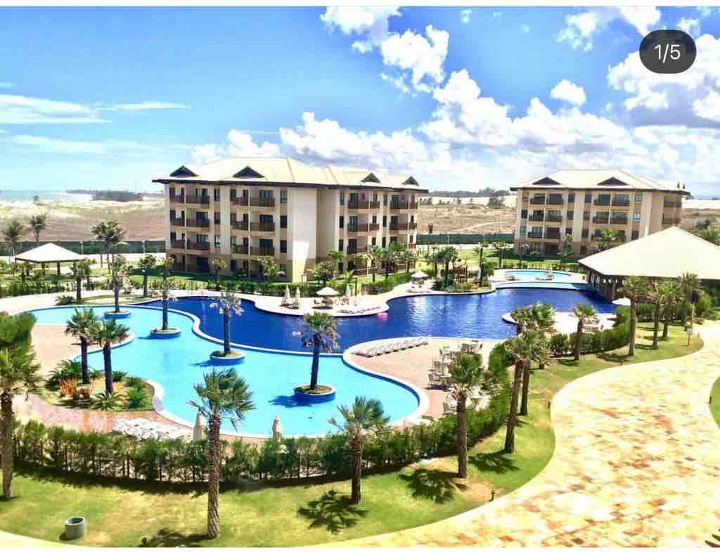 A tranquil outdoor area features a large swimming pool surrounded by palm trees, with lounge chairs positioned along the water's edge. Nearby, a shaded gazebo provides a relaxed seating area. The backdrop includes multiple two-story buildings and a clear blue sky with scattered clouds.