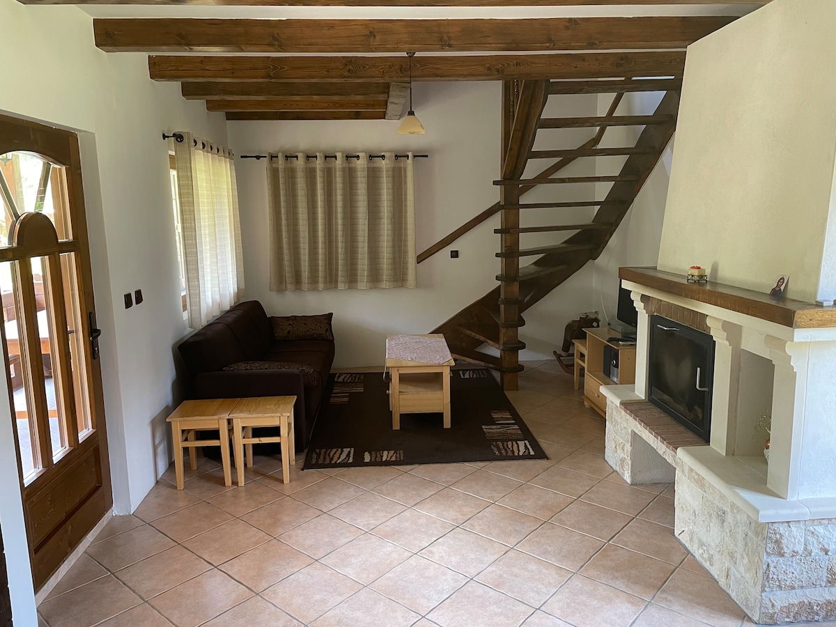 A spacious living area features a dark sofa paired with two light-colored wooden tables, set on a neutral-toned rug. Exposed wooden beams and tiled flooring contribute to a rustic ambiance. A staircase ascends to the upper level, while a fireplace is positioned beside a media unit.