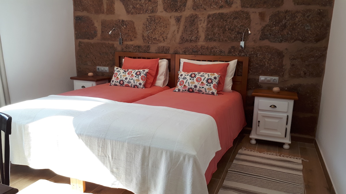 A well-lit sleeping area features two individual beds, each dressed in coral-colored linens. Decorative pillows add a touch of color to the beds. The room is accented by a natural stone wall, with side tables on either side and warm wooden flooring throughout.