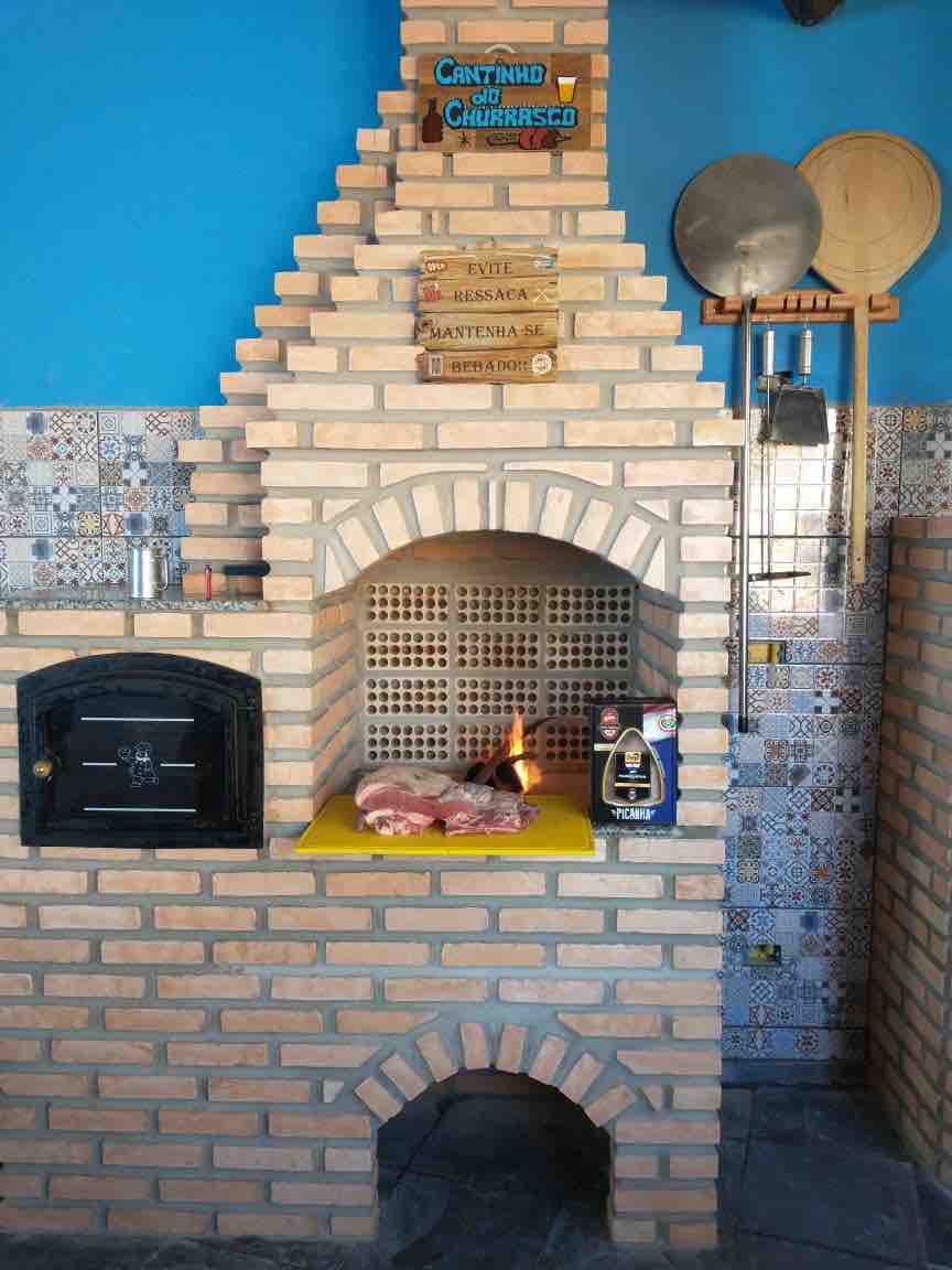 A brick barbecue grill is centrally positioned, featuring an open flame and a yellow cutting board with raw meat. The surrounding area displays blue walls and patterned tiles, with cooking utensils hanging on the wall, creating an organized space for outdoor cooking.
