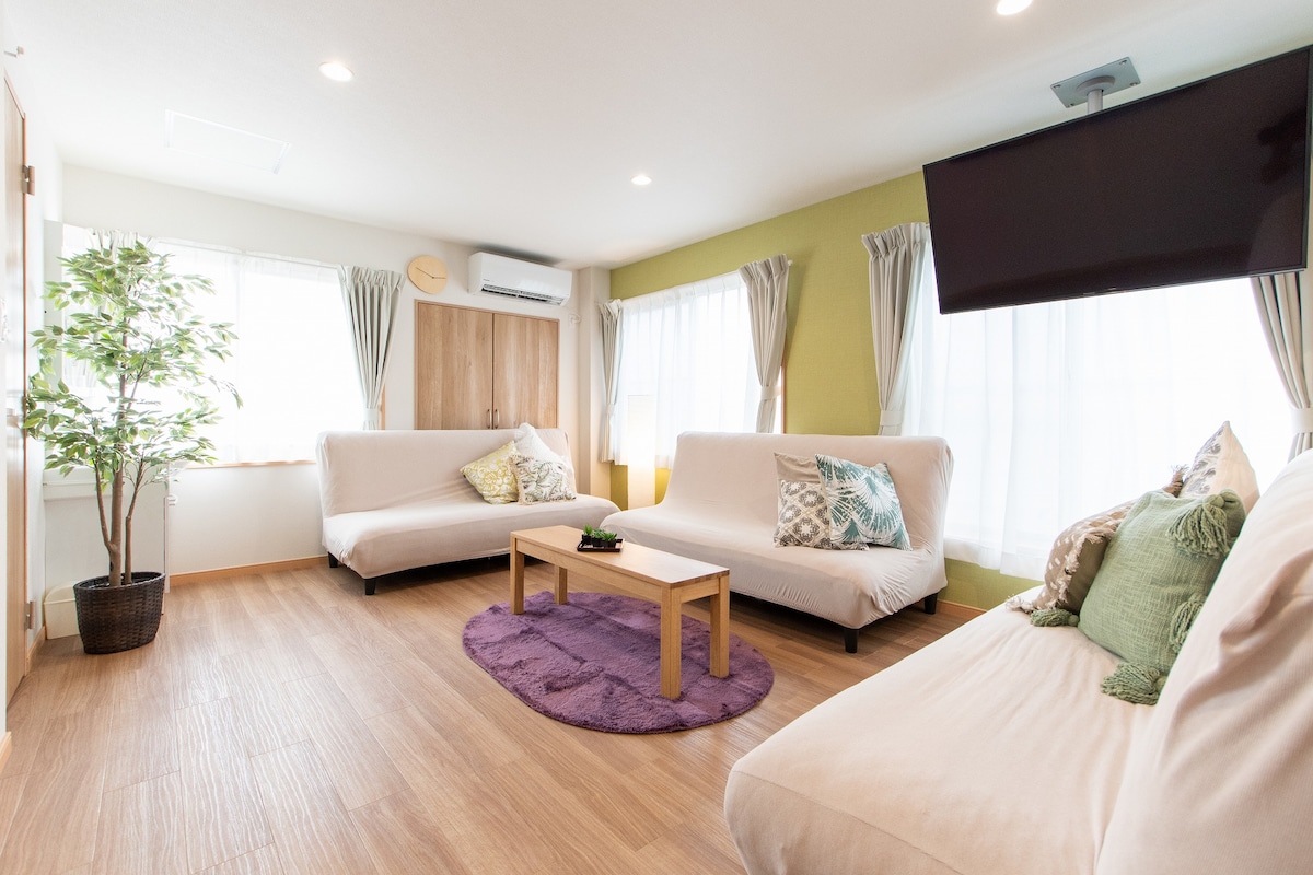 A bright living area features two spacious sofa beds and a coffee table placed on a plush purple rug. Natural light fills the space through large windows dressed with light curtains, complemented by indoor plants and a wall-mounted television.