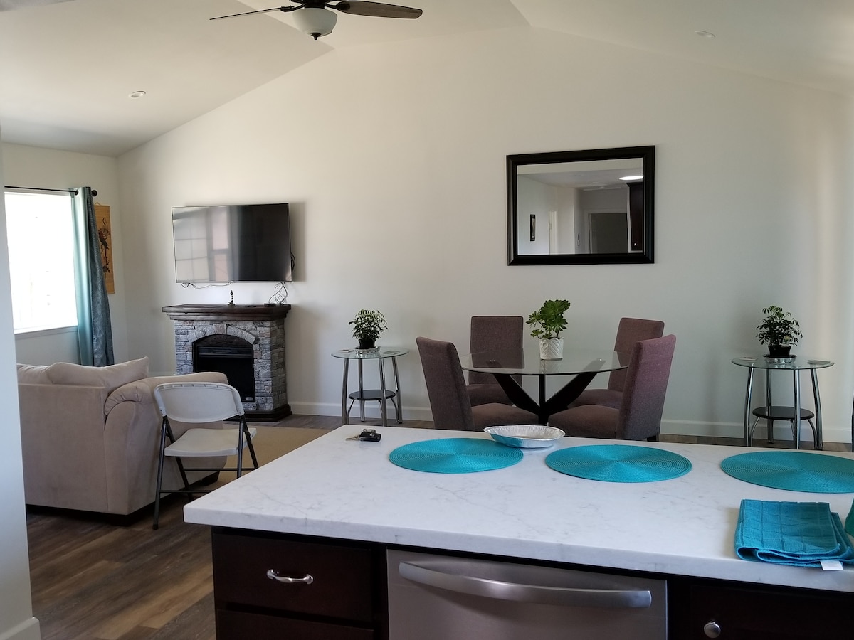 An open living space features a cozy seating area with a light-colored sofa and two side chairs. A round dining table with four chairs is positioned nearby, accompanied by small glass-top tables. A mounted television and decorative plants enhance the inviting environment.