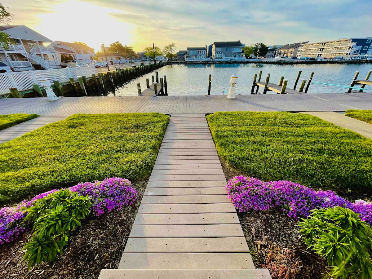 A wooden boardwalk leads through lush green grass and vibrant purple flowers towards a serene bay. Soft sunlight illuminates the scene, highlighting the peaceful water and nearby buildings. The setting offers a tranquil experience by the water's edge.