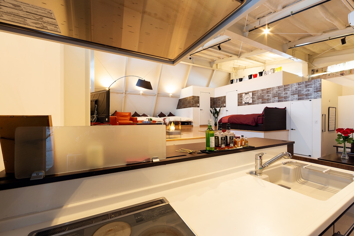 The kitchen area is equipped with a stove and countertop, offering a view into the spacious living area. The high ceilings and creative lighting highlight an open layout, complemented by various bottle placements on the countertop, enhancing the overall aesthetic.