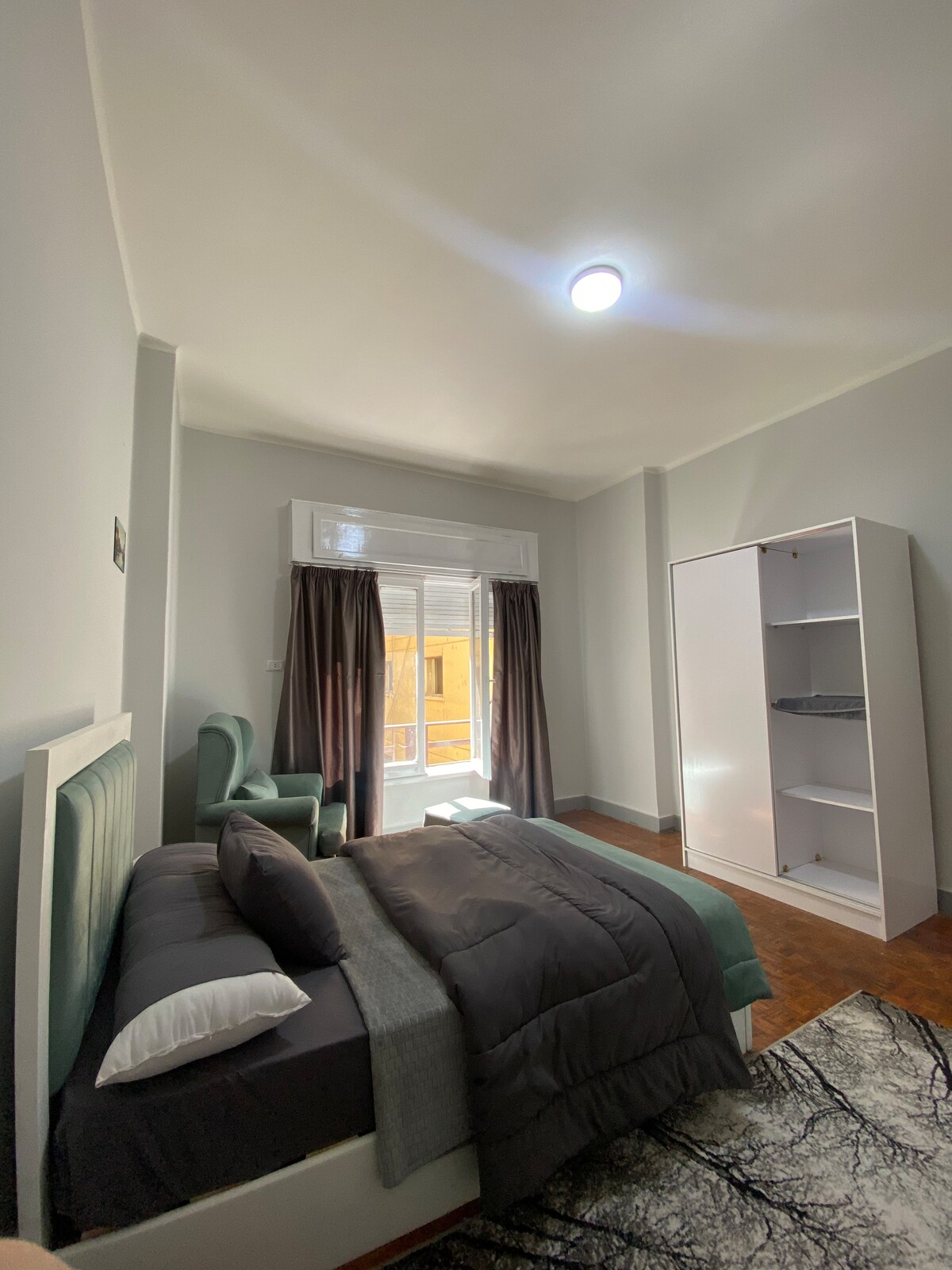 A spacious single room features a comfortable bed layered with dark bedding and pillows. Natural light enters through large windows, partially covered by dark curtains. A cozy chair sits beside the bed, and a white wardrobe is positioned against the wall, contributing to the inviting atmosphere.