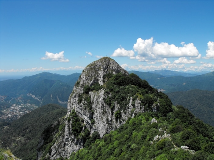 A large rocky peak is surrounded by lush greenery, rising prominently against a clear blue sky. The distant mountains add depth to the scene, while scattered clouds float gently above, enhancing the natural beauty of the landscape.