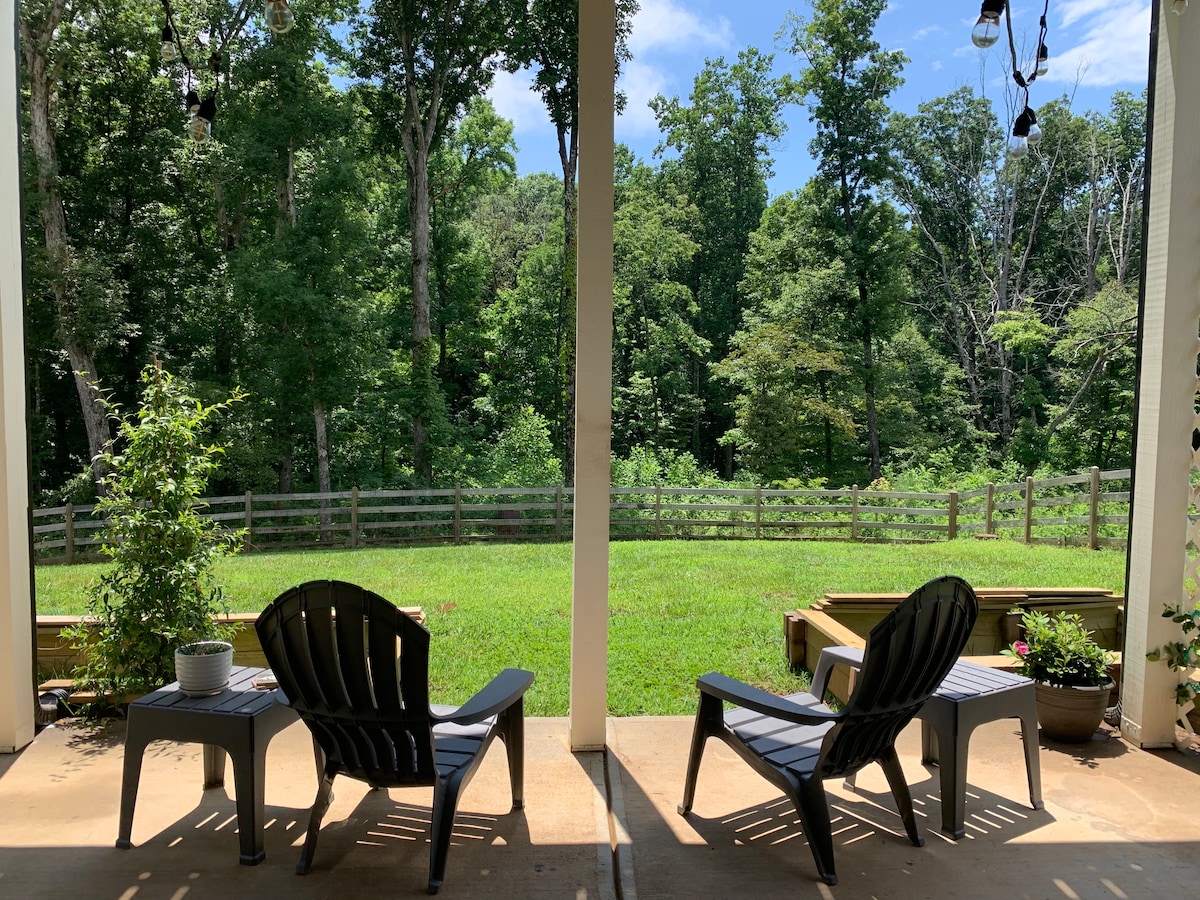 Two sleek chairs are positioned on a covered patio, providing a view of an expansive green yard and a wooded area. A wooden fence borders the property, and a few potted plants add a touch of nature to the scene.