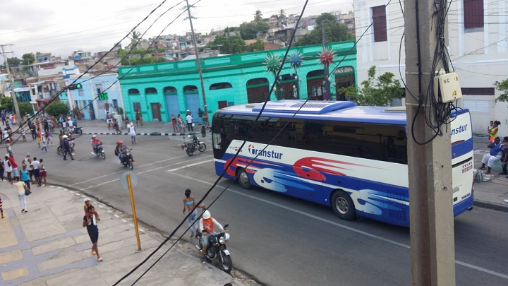 A busy street scene is depicted, featuring a blue and white bus moving down the road. Alongside the bus, motorcycles and pedestrians are visible, highlighting the area's vibrant activity. Colorful buildings, including a green structure, create an engaging urban backdrop.