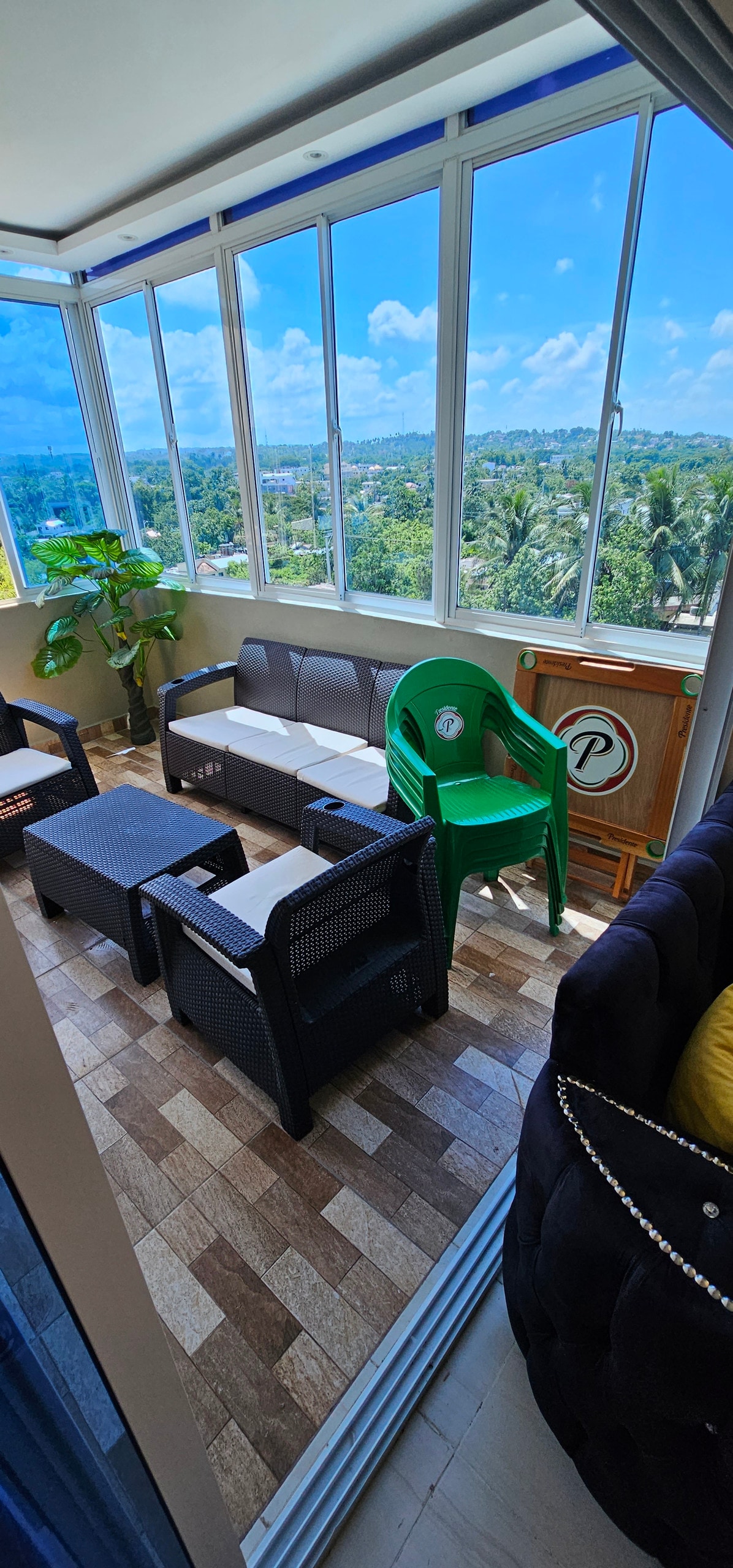 A sunlit seating area features a mix of seating options, including a dark sofa and several armchairs, with a view of the surrounding greenery through large windows. A potted plant adds a touch of nature, complementing the relaxing atmosphere.