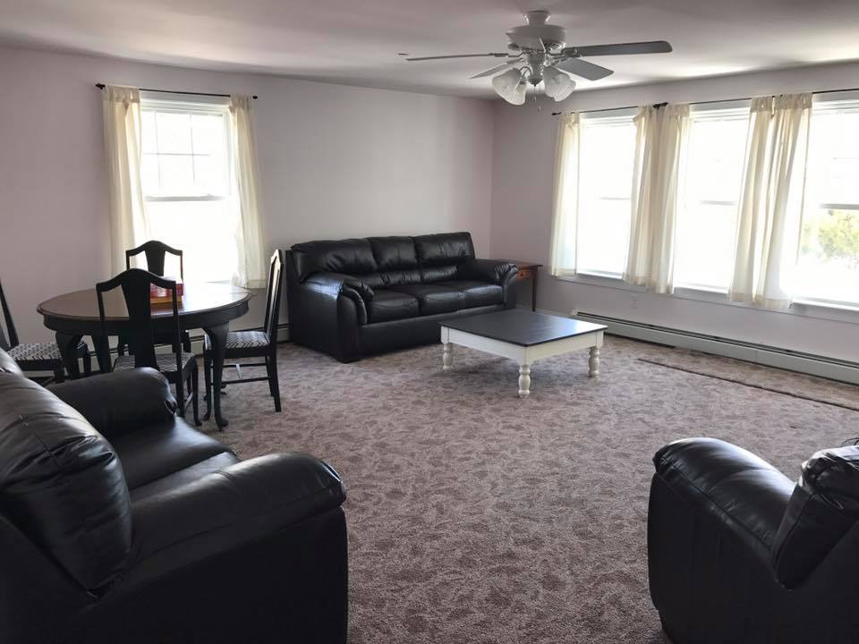 A spacious living area features two dark leather sofas and a comfortable armchair arranged around a coffee table. A dining table with chairs sits in one corner. Natural light filters through three large windows adorned with light curtains, illuminating the carpeted floor.