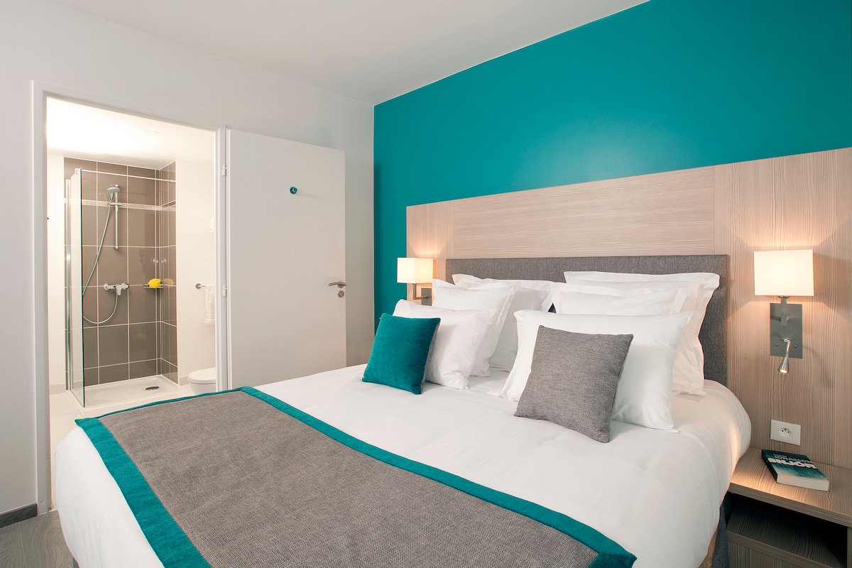 A well-appointed sleeping area features a queen-sized bed with decorative pillows against a turquoise accent wall. Soft lighting from mounted lamps enhances the ambiance. A bathroom is visible in the background, showcasing modern fixtures and a glass-enclosed shower.