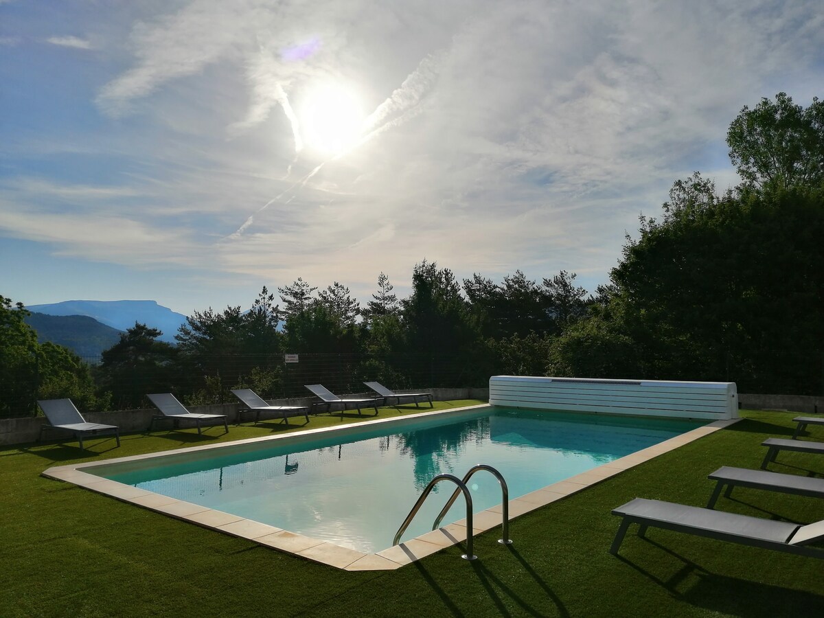 An outdoor pool surrounded by manicured grass features lounge chairs positioned along its perimeter. The tranquil water reflects the sky, with soft clouds drifting above. A backdrop of trees and hills creates a serene setting, illuminated by the morning sun.