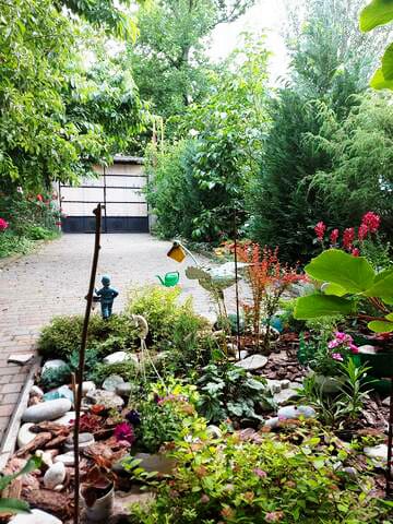 A green and serene garden area is highlighted, featuring various plants and shrubs along a stone pathway. Decorative elements include small sculptures and colorful flowers, enhancing the natural setting. Tall trees provide shade, creating a peaceful atmosphere for relaxation.