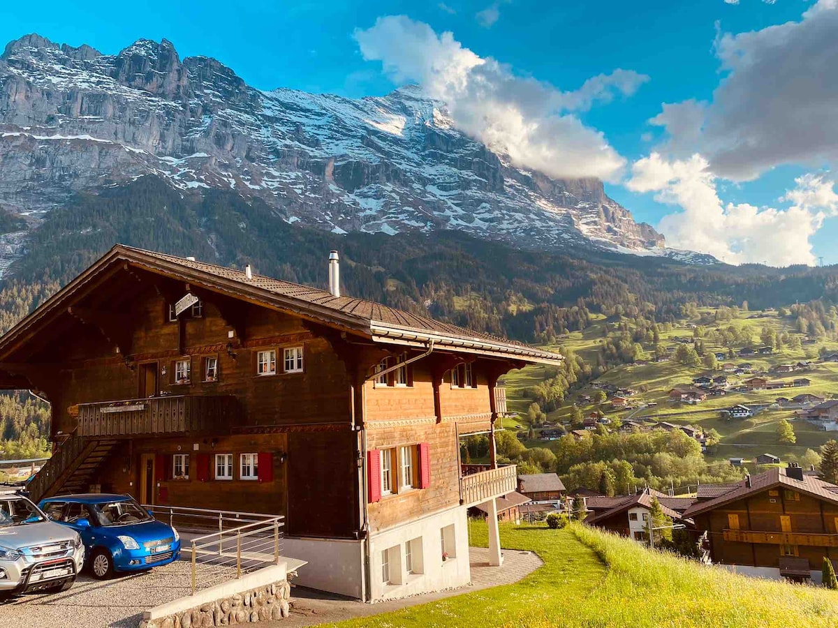 A wooden chalet is nestled against a backdrop of mountains, showcasing snow-capped peaks and lush green valleys. The structure features multiple levels, with balconies offering outdoor space, while parked cars are visible on a gravel driveway. The scene is illuminated by natural light from a clear sky.