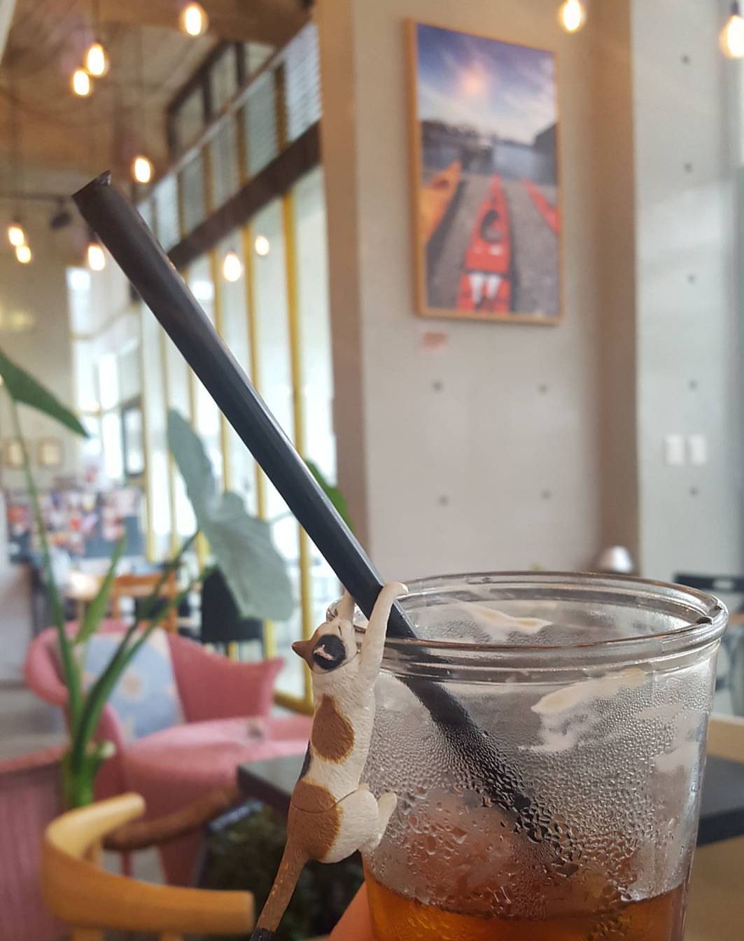 A glass of iced tea is seen with a playful toy figure clinging to its straw. The background features a modern café setting with warm lighting, colorful seating, and a large artwork hanging on the wall, creating a lively atmosphere.