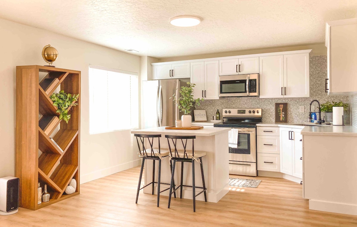 A modern kitchen is showcased with white cabinetry and stainless steel appliances. A wooden bookshelf with geometric design is filled with decorative items. Two black bar stools are positioned at a central island, while natural light filters through a nearby window.