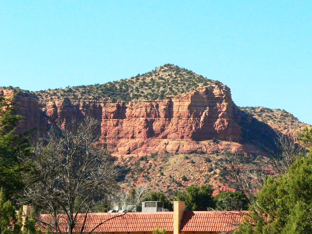 A view showcasing the striking contours of a red rock formation against a clear blue sky. The rocky landscape features patches of greenery and low shrubs at its base, highlighting the natural beauty of the surroundings.