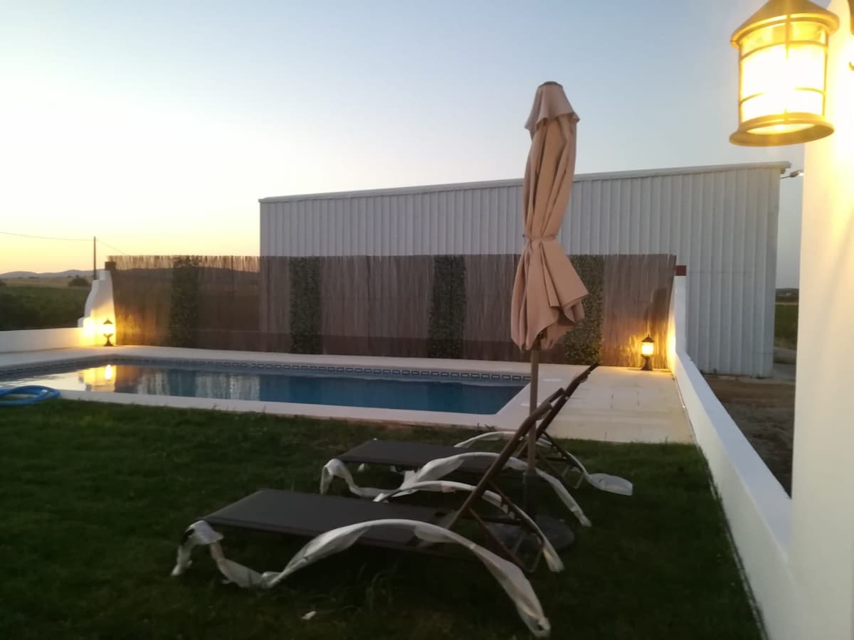 A tranquil outdoor space features a swimming pool surrounded by a well-maintained lawn. Two lounge chairs are positioned beside the pool, and a sun umbrella is raised for shade. Soft lighting from a wall lamp creates an inviting ambiance as dusk approaches.