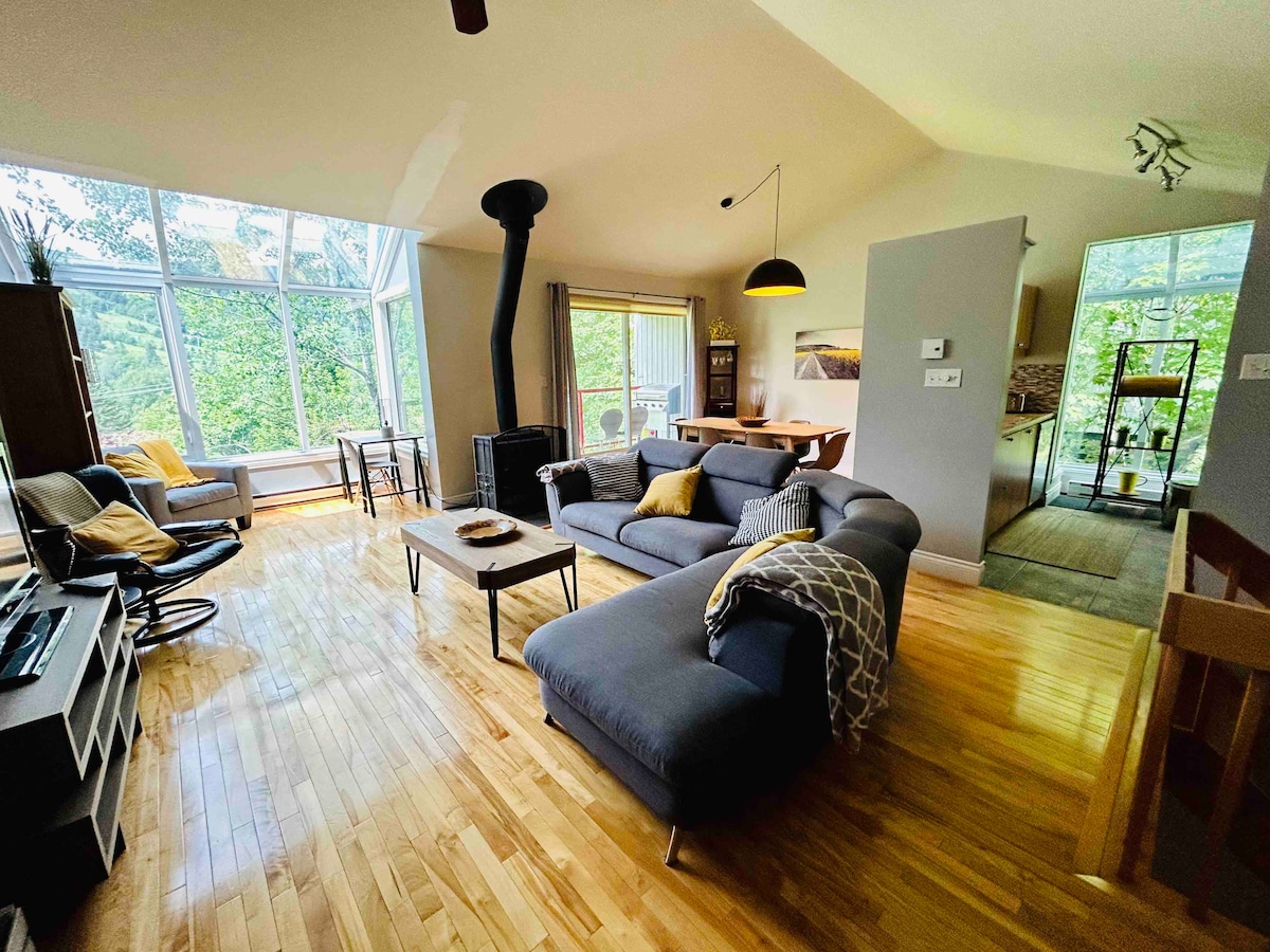 An open-concept living area is illuminated by large windows, allowing natural light to fill the space. A cozy sectional sofa and armchair offer seating options, while a wooden table is set for two. A sleek fireplace serves as a focal point within the room.