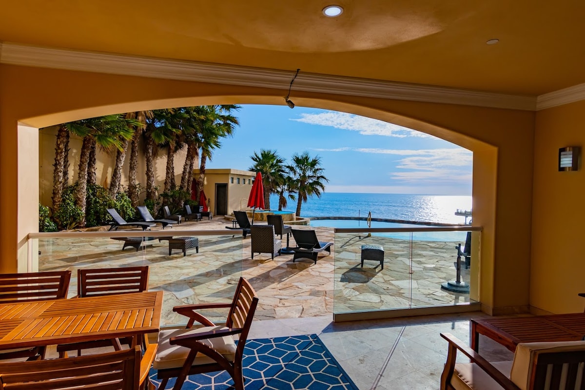 A shaded outdoor terrace offers a view of the ocean, highlighted by palm trees and deck chairs. The ocean sparkles in the distance, with a bright sky above. A wooden dining table and blue rug frame the scene, inviting relaxation near the water.