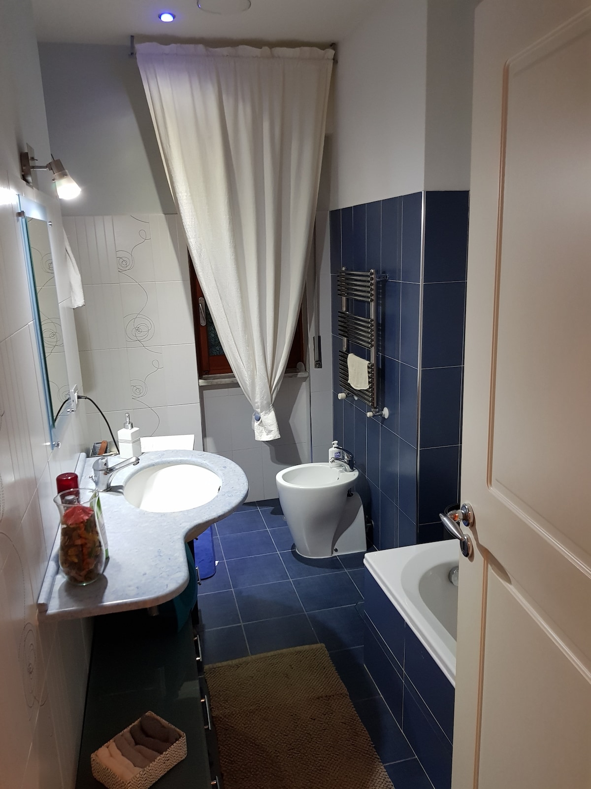 A well-organized bathroom features a curved countertop with dual sinks and a large mirror above. A bathtub is positioned next to a wall-mounted towel rack. Blue tiles provide a calm color palette, complemented by a window covered with a flowing white curtain, allowing natural light to enter.