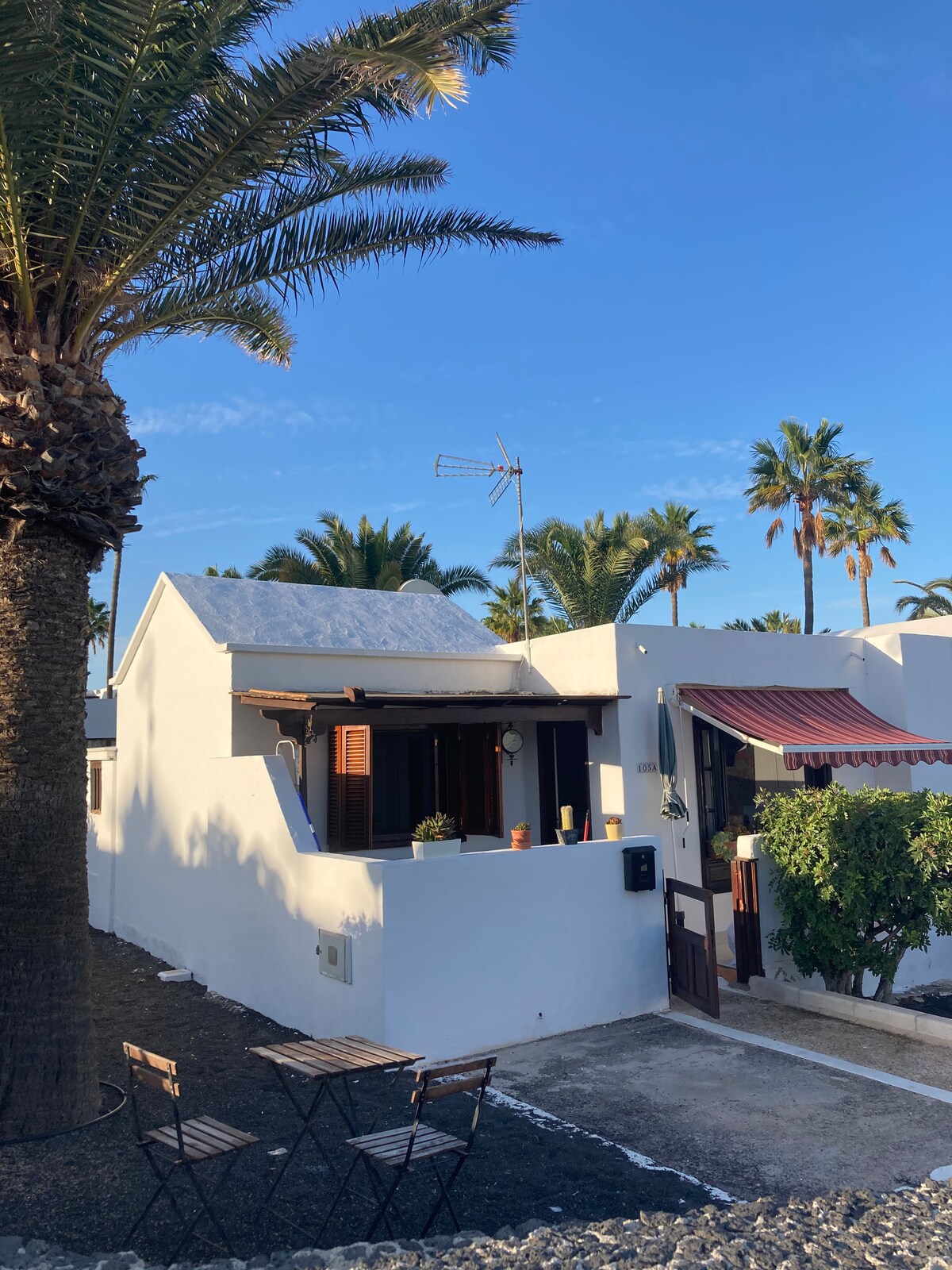 A white bungalow is surrounded by palm trees, featuring a small outdoor seating area with a table and chairs. The facade has wooden shutters and a decorative awning, while a clear blue sky enhances the tropical ambiance.