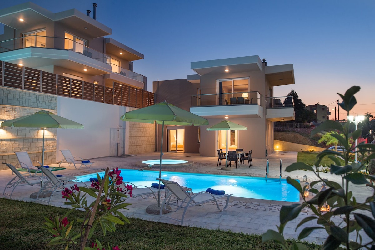 Two modern villas are depicted, each featuring a private pool and spacious outdoor area. Lounge chairs are positioned beside the pool, accompanied by umbrellas for shade. A Jacuzzi can be seen integrated into the pool area, with the villas illuminated under a twilight sky.