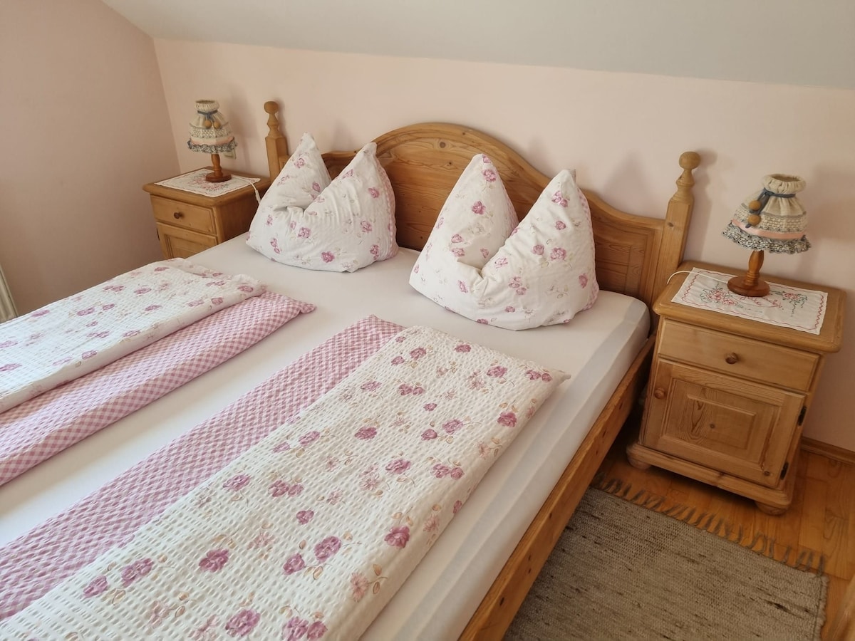The sleeping area features a spacious bed dressed with floral-patterned linens and plush pillows. Wooden nightstands, each with a decorative lamp, are positioned beside the bed. Soft light enhances the cozy ambiance of the room, accented by light pink walls.
